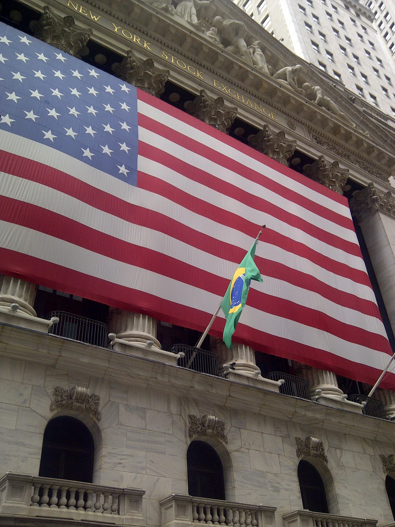 The New York Stock Exchange facade covered by a monumental United States flag, with a smaller Brazilian flag below.