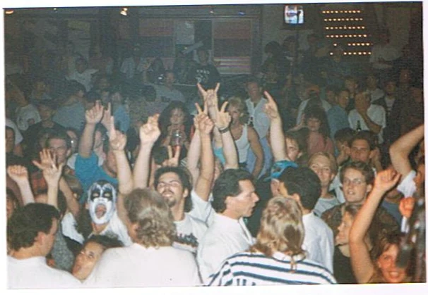 A crowded room with a fan in KISS makeup raising both hands.